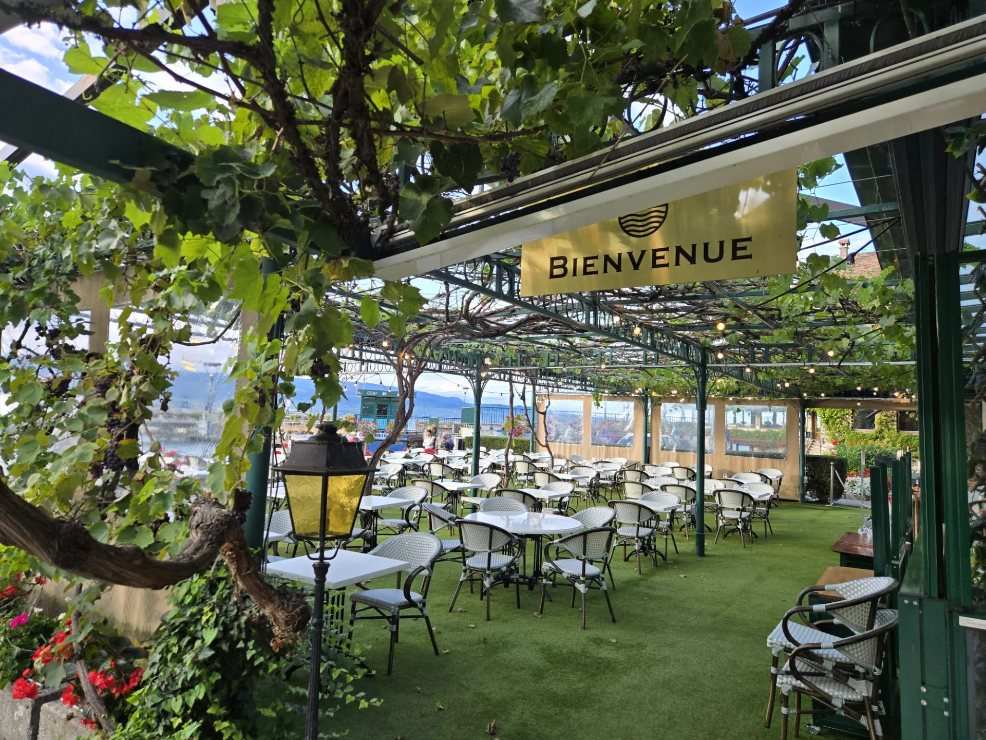 Terrasse du restaurant sous une treille de vigne avec panneau Bienvenue et chaises bistrot
